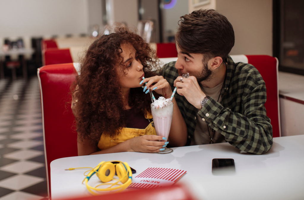 A male and female sitting in a restaurant sipping a milkshake from the same cup.
